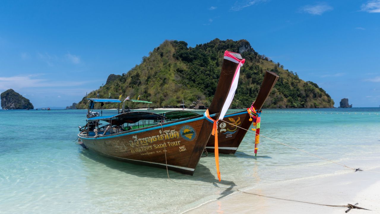 Two traditional longtail boats docked on a pristine tropical beach against a backdrop of lush islands.